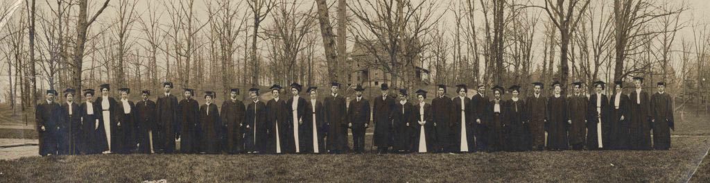 The graduating class of 1909 in black caps and gowns standing in a single file on the campus grounds.