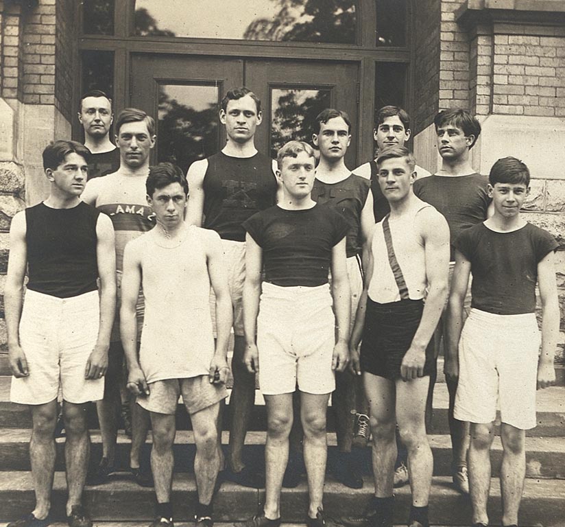 Eleven men from the track and field team, dressed in track shorts and shirts, stand on the steps of Bowen Hall.