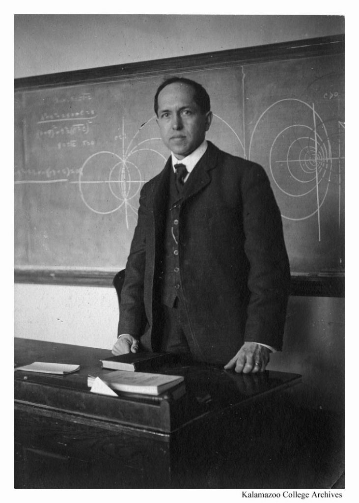 Clark Benedict Williams in a suit and tie stands behind his desk in front of the blackboard.